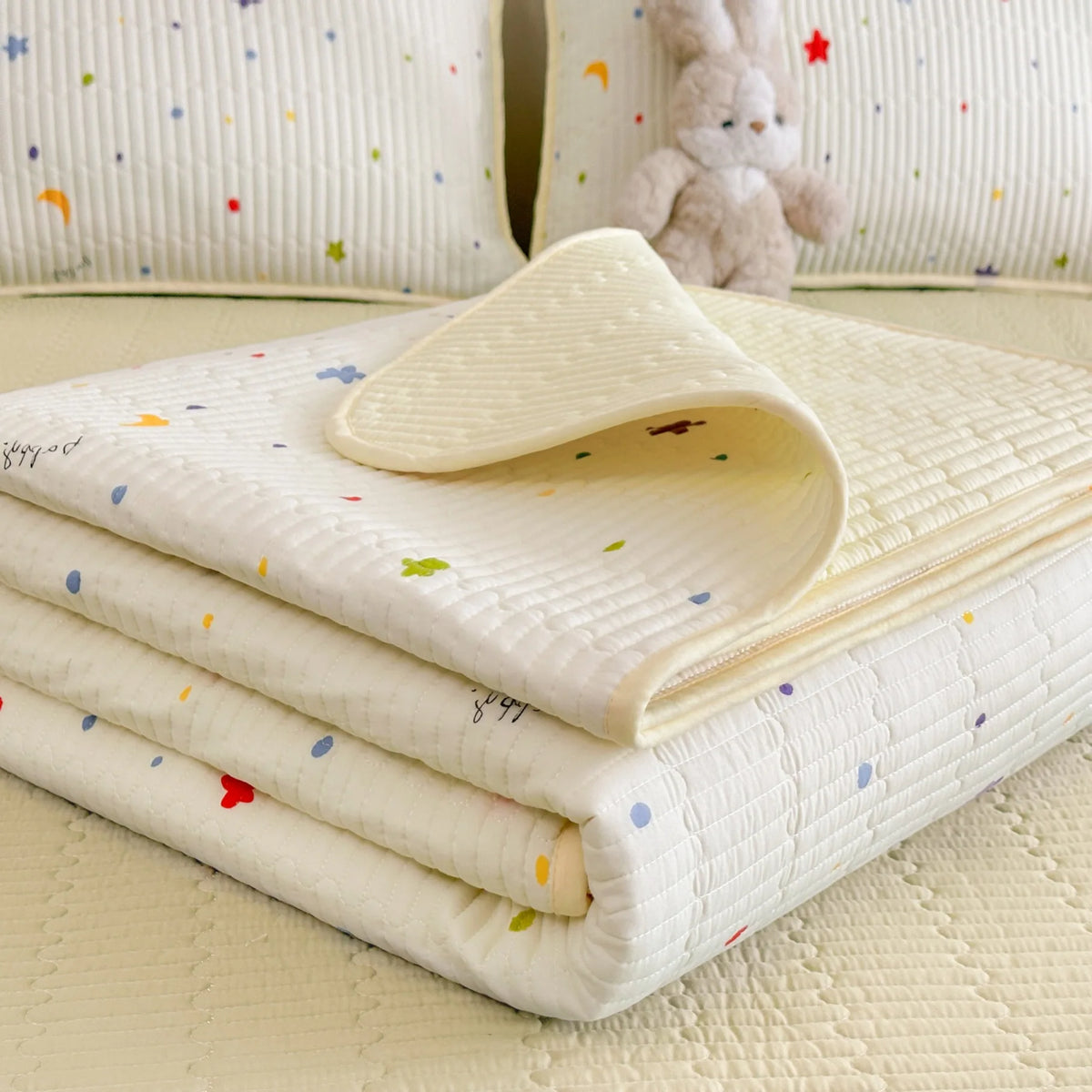 A stack of white quilted fabric and a folded cream cloth with a small rabbit toy in the background.