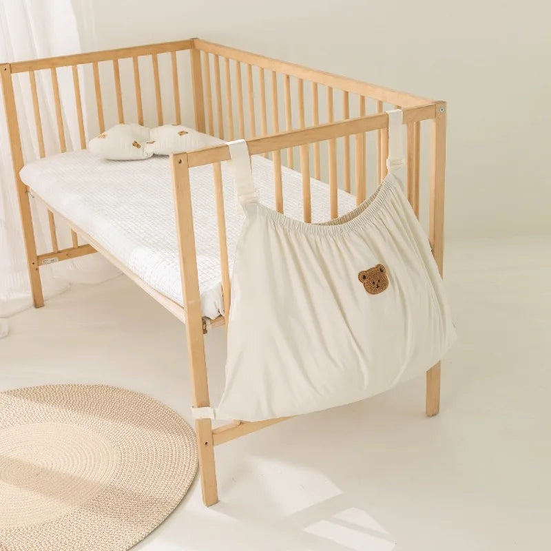 A wooden crib with a white mattress and bedding, featuring an adorable bear-themed storage bag.