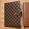 A black and brown checkered PU leather portfolio with a leather strap resting on a tan leather surface.