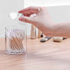 A hand is opening a clear storage jar with a lid, revealing numerous white cotton swabs inside.