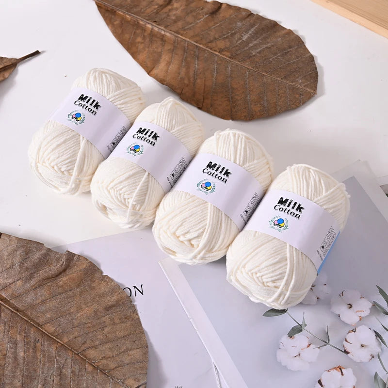 Several balls of white milk cotton yarn are displayed next to a large dried leaf on a white surface.