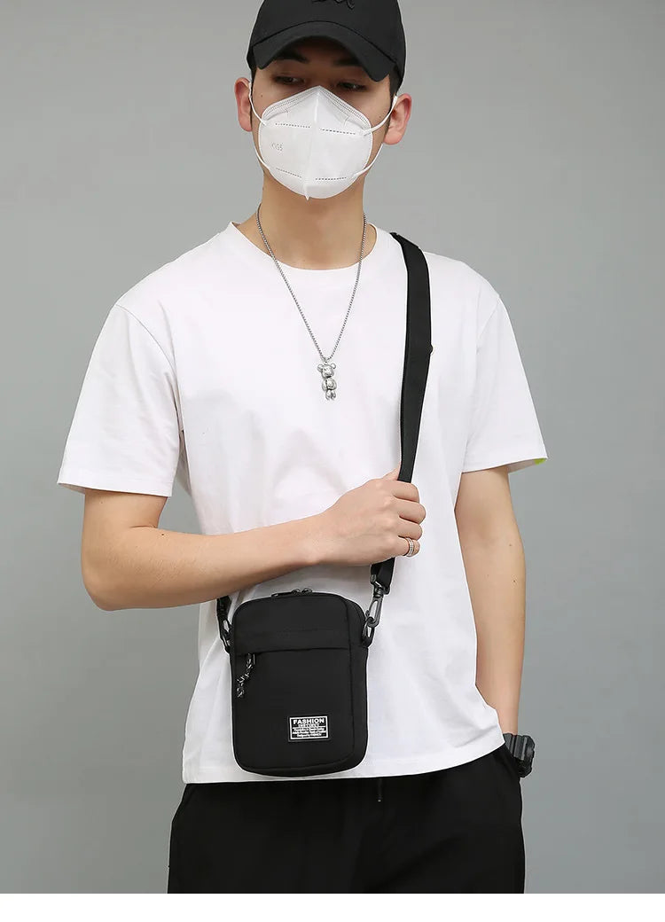 A person wearing a white t-shirt and black cap holds a black mini crossbody phone bag with an adjustable strap.