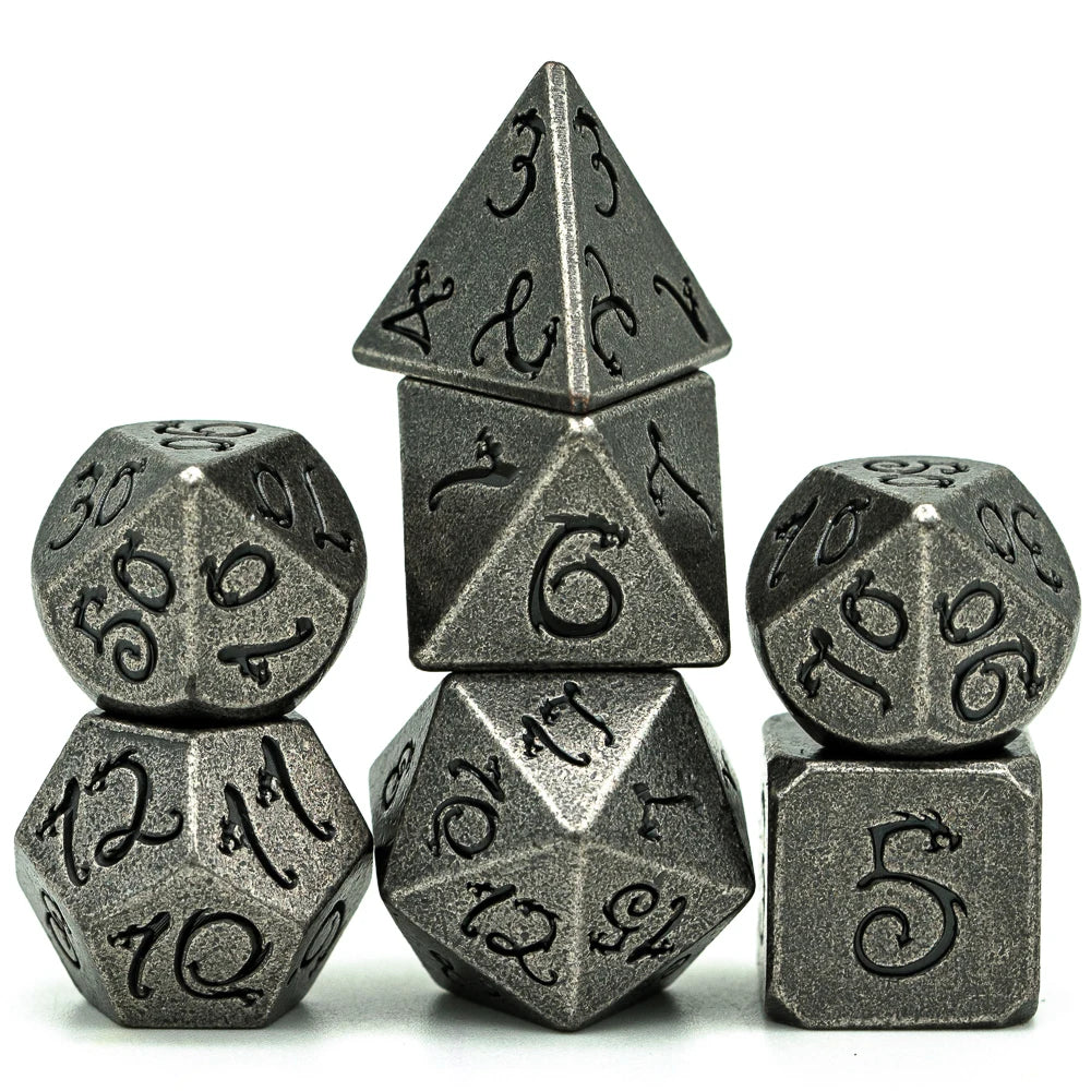 A set of seven metal polyhedral dice with a dragon font design stacked and scattered on a white background.