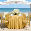 A round table set for an outdoor event with a golden satin tablecloth, white flowers, and gold chairs by the sea.