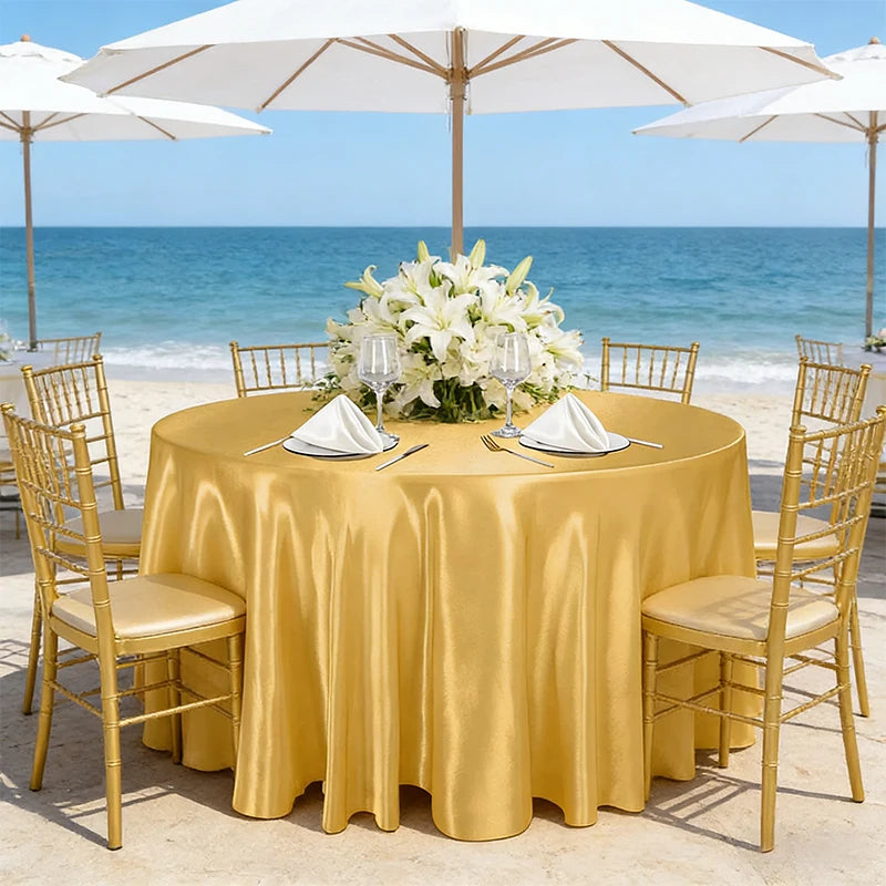 A round table set for an outdoor event with a golden satin tablecloth, white flowers, and gold chairs by the sea.