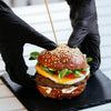 Hands in black gloves assemble a gourmet burger on a black slate board.
