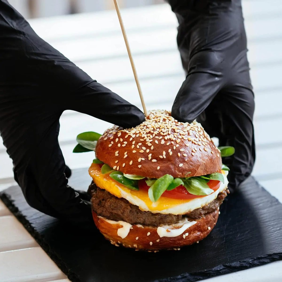 Hands in black gloves assemble a gourmet burger on a black slate board.