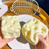 Eine Hand hält ein gelbes, weiches Käse-Knetspielzeug auf einem weißen Teller neben einem Drahtgeflecht-Werkzeug.