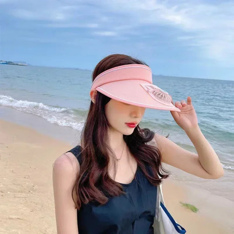 A woman wearing a pink USB rechargeable cooling fan hat stands on a beach with the ocean in the background.