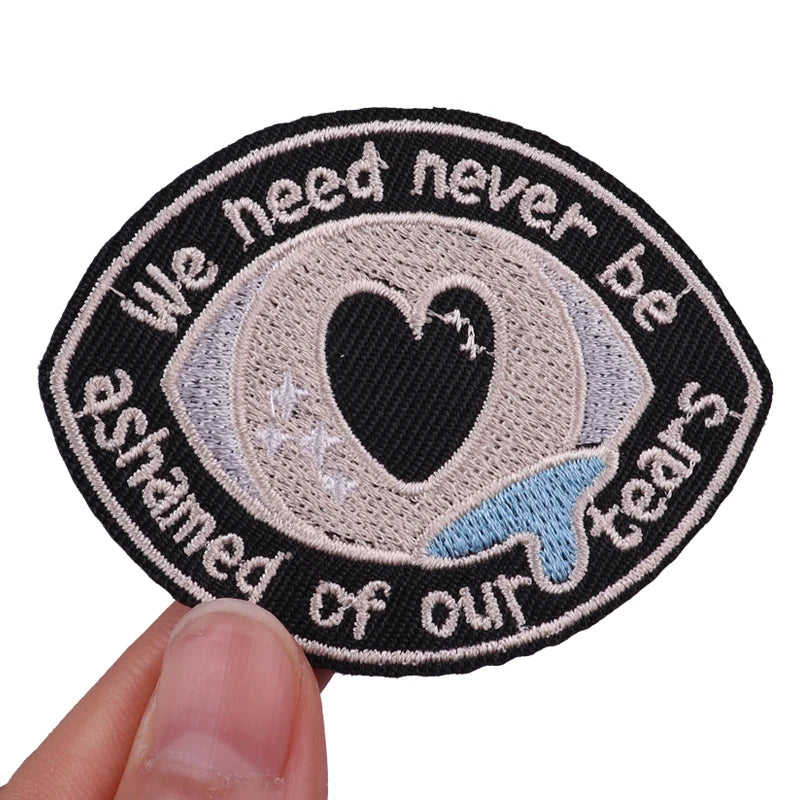 Black embroidered patch with white text and a heart design that reads We need never be ashamed of our tears