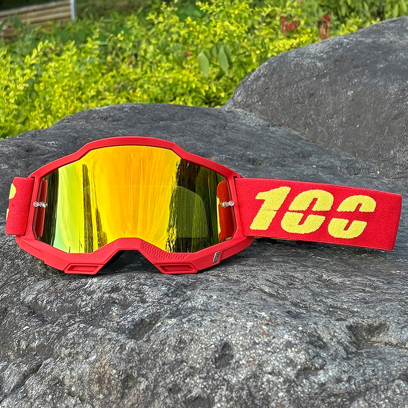 Red motocross goggles with yellow mirrored lenses resting on a dark gray rock.