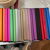 A row of colorful trading card protective display cases with various colored spines arranged horizontally.