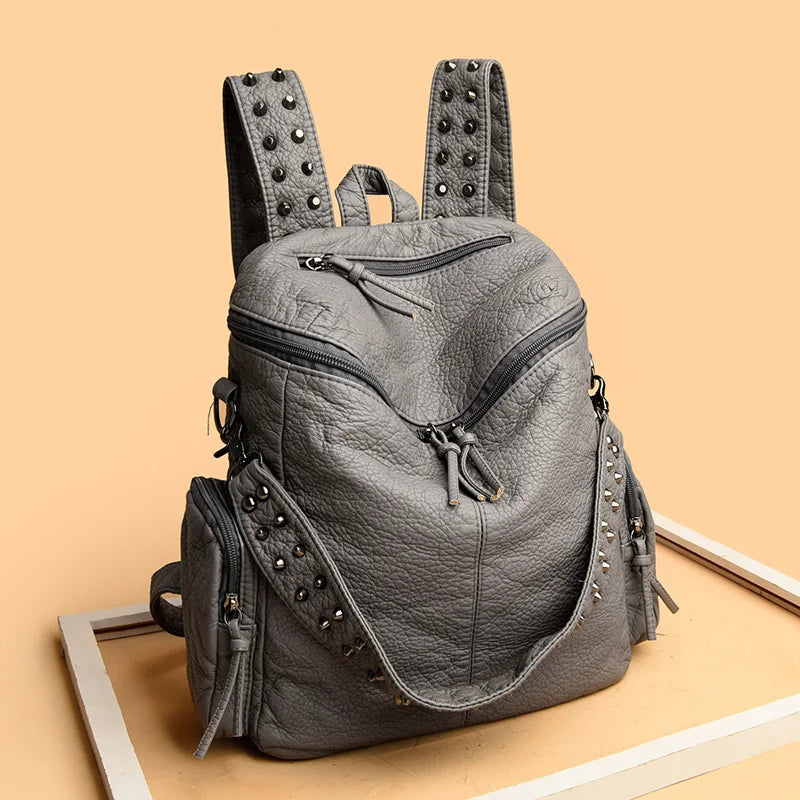 A gray soft leather backpack with metal rivets and a flap closure is displayed on a light wooden surface.
