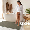 A woman sits in a white bathtub surrounded by natural decor, with a green waffle weave bath mat on the floor.