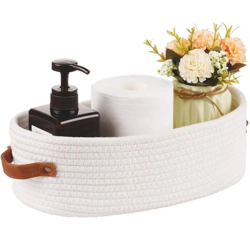A white woven cotton rope storage basket holding a roll of toilet paper and a floral arrangement.