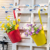 Yellow and red pots filled with flowers and dragonfly clips hanging on a white wooden fence.