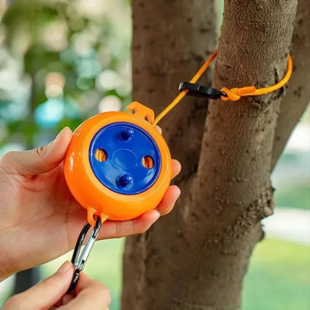 A hand holds an orange retractable clothesline with a blue disc and black strap attached to a tree trunk.