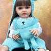 A lifelike silicone baby doll wearing a blue bear hat and a matching blue knitted outfit.