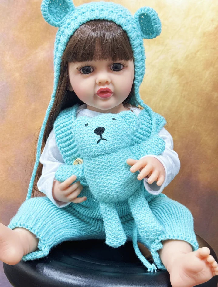 A lifelike silicone baby doll wearing a blue bear hat and a matching blue knitted outfit.