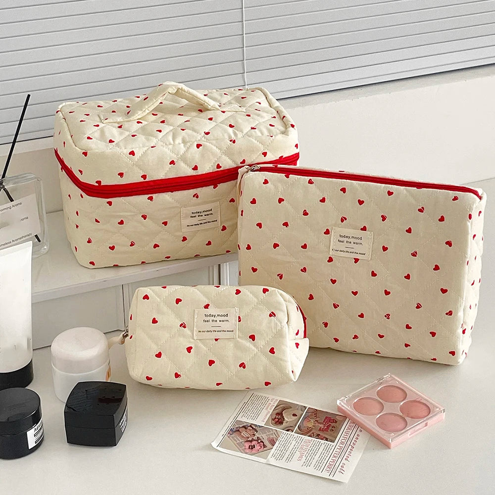 A quilted white makeup bag with a red trim and heart pattern is shown next to various makeup products.