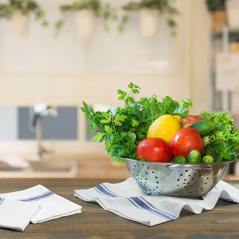 A bowl of fresh vegetables including tomatoes, lemons, and leafy greens placed on a wooden table with striped kitchen towels.