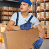 A young man wearing blue work gloves and a blue cap is handling a cardboard box in a warehouse setting.