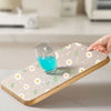 A hand places a glass of blue liquid onto a wooden drawer mat decorated with white and green daisy flowers.