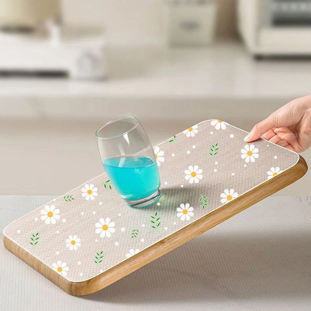 A hand places a glass of blue liquid onto a wooden drawer mat decorated with white and green daisy flowers.