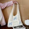 A handwoven knit tote bag in off-white with a hollow design is being held by a person against a wooden background.
