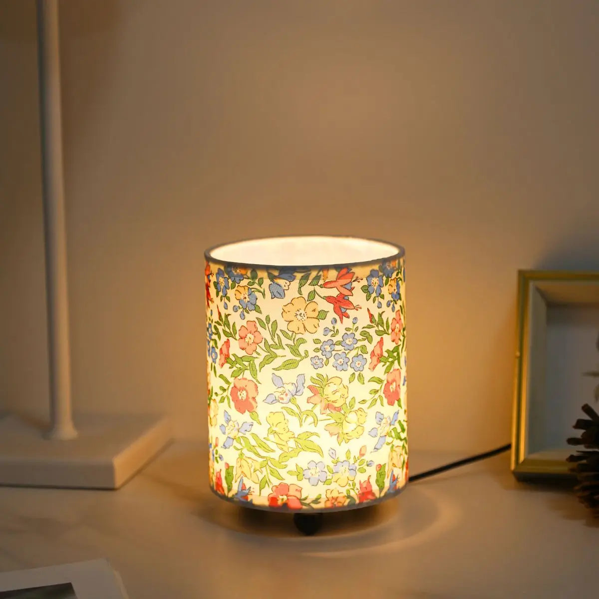 A cylindrical desk lamp with a floral fabric shade emits a warm glow on a white surface.