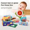 A baby is interacting with a colorful sensory tissue box filled with crinkle scarves and fabric toys.