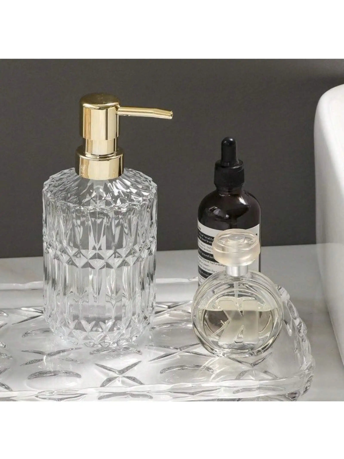 A clear glass soap dispenser with a gold pump and a small glass bottle are displayed on a clear tray.