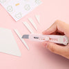 Eine Hand hält ein rosa Mini-Klaue-Werkzeugmesser mit Klinge neben weißen Papierfetzen.