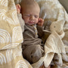 A newborn baby is sleeping in a light yellow and beige patterned fabric, wearing a brown outfit.
