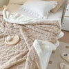 A beige double-layer fleece throw blanket draped over a white bed with a white pillow.