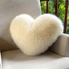 A fluffy, heart-shaped cream-colored faux fur throw pillow resting on a light-colored sofa near a window.
