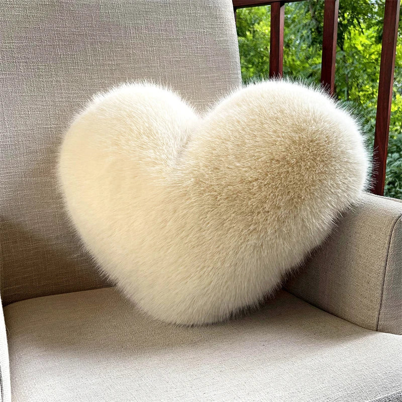A fluffy, heart-shaped cream-colored faux fur throw pillow resting on a light-colored sofa near a window.