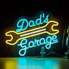 Neon sign spelling out Dad's Garage in bright blue and yellow outline on a dark wooden background.