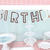Dekorativer Banner mit 'birthday' auf blauer Wand, weiß und roségold und gestapelte weiße Plastikdeko