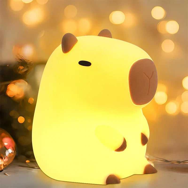 A yellow silicone night light shaped like a cow, glowing softly against a blurred background of warm bokeh lights.
