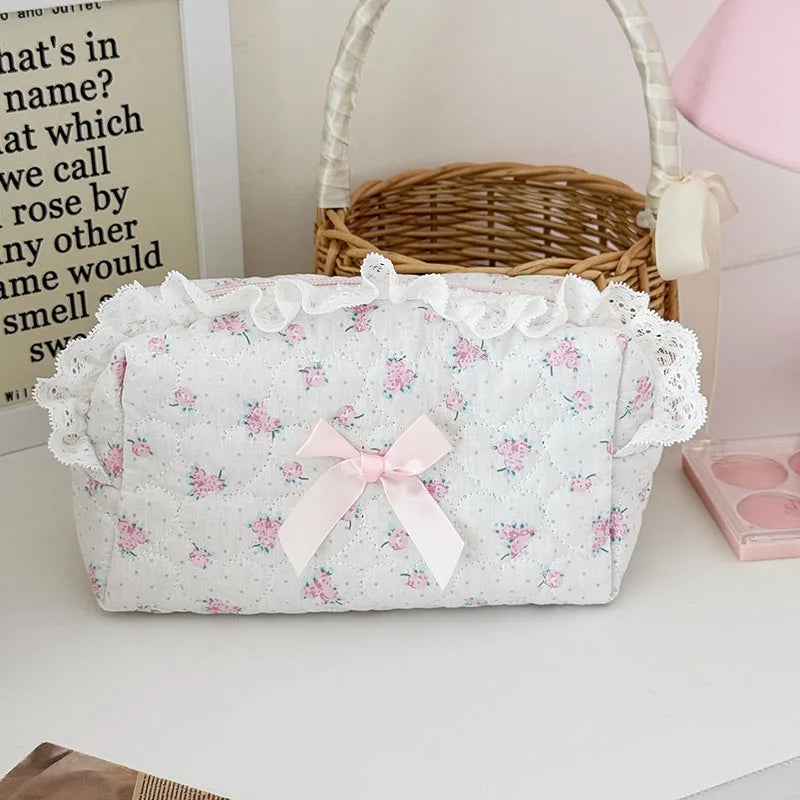 A white cosmetic pouch with a vintage rose print and delicate lace trim is displayed next to a woven basket.