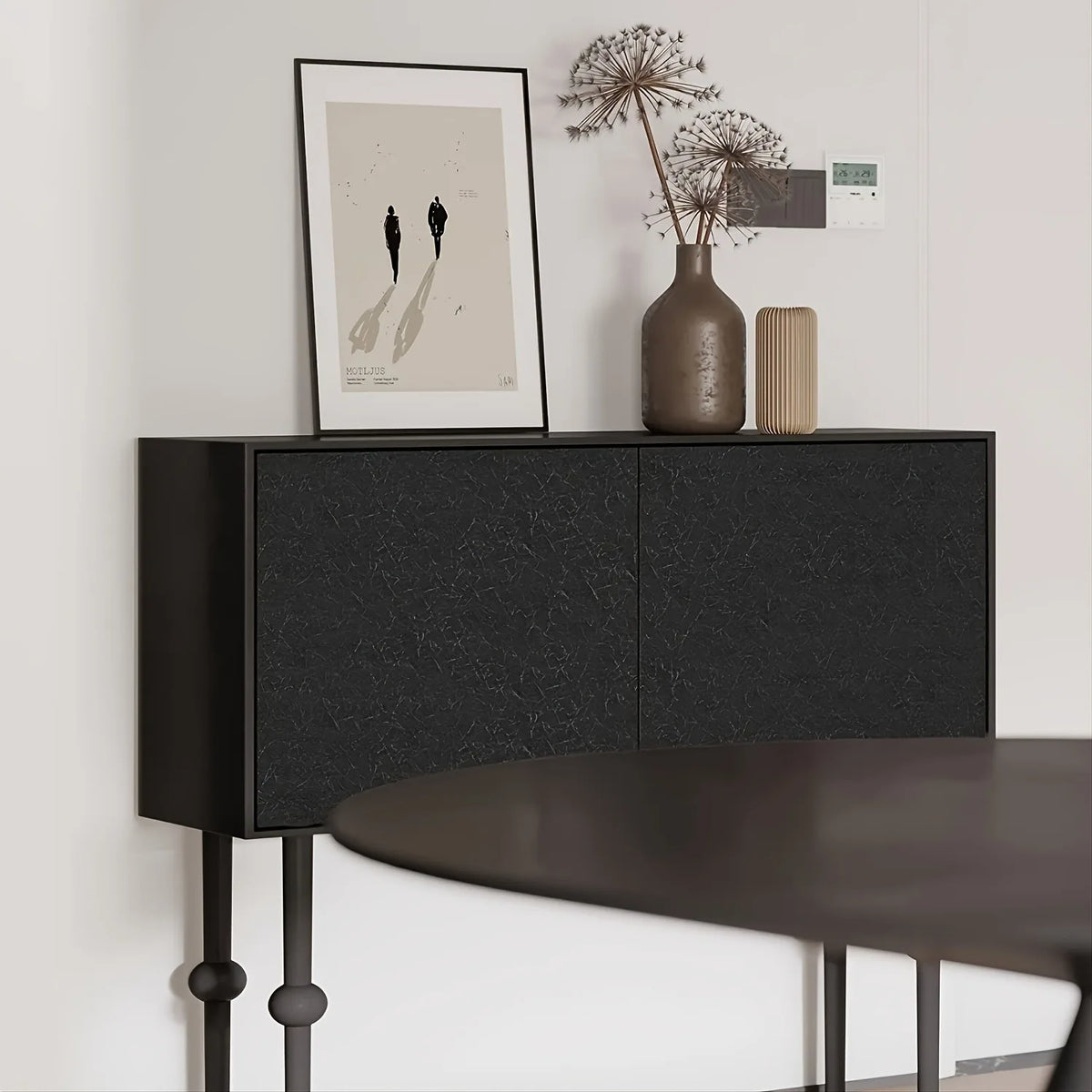 A dark wood cabinet with a textured black front holds a framed picture and a dried plant arrangement.