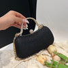 A black shimmering envelope clutch with a gold chain strap is held by a hand on a white fabric surface.