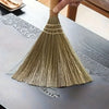 A hand is holding a natural grass broom with a woven handle, resting it on a wooden table surface.