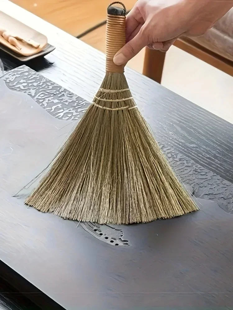 A hand is holding a natural grass broom with a woven handle, resting it on a wooden table surface.