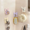 Un juego de ganchos decorativos de mariposa giratoria en rosa, morado y crema montados en una pared beige clara.