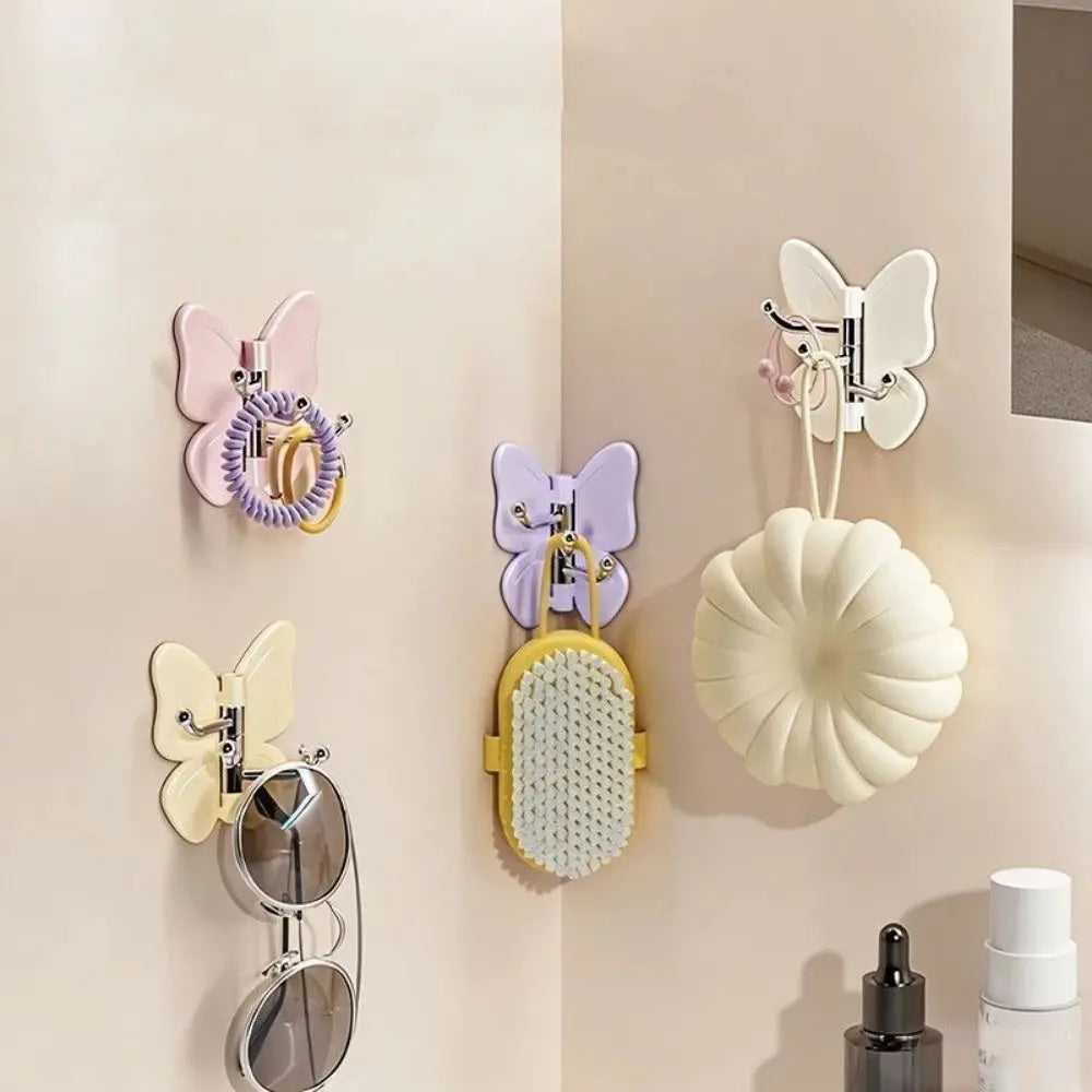 Un juego de ganchos decorativos de mariposa giratoria en rosa, morado y crema montados en una pared beige clara.