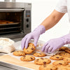 A hand wearing light purple disposable nitrile gloves is placing round cookies onto a baking sheet.