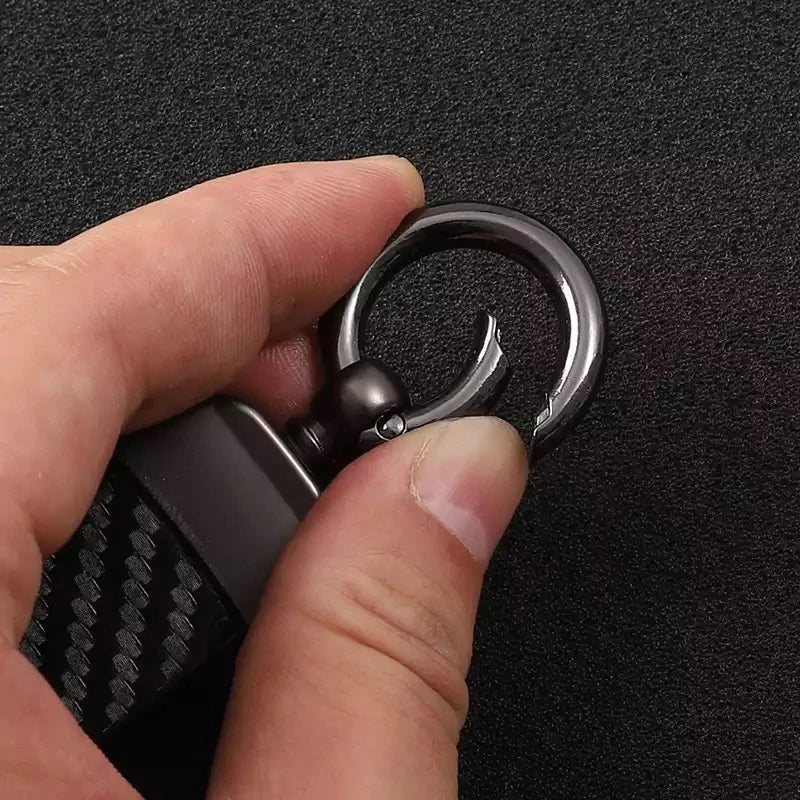A hand holds a black carbon fiber style leather keychain with a silver swivel buckle.