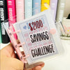 Eine Hand hält einen klaren Ordner mit der Aufschrift 'Savings Challenge' und Dollarbeträgen.